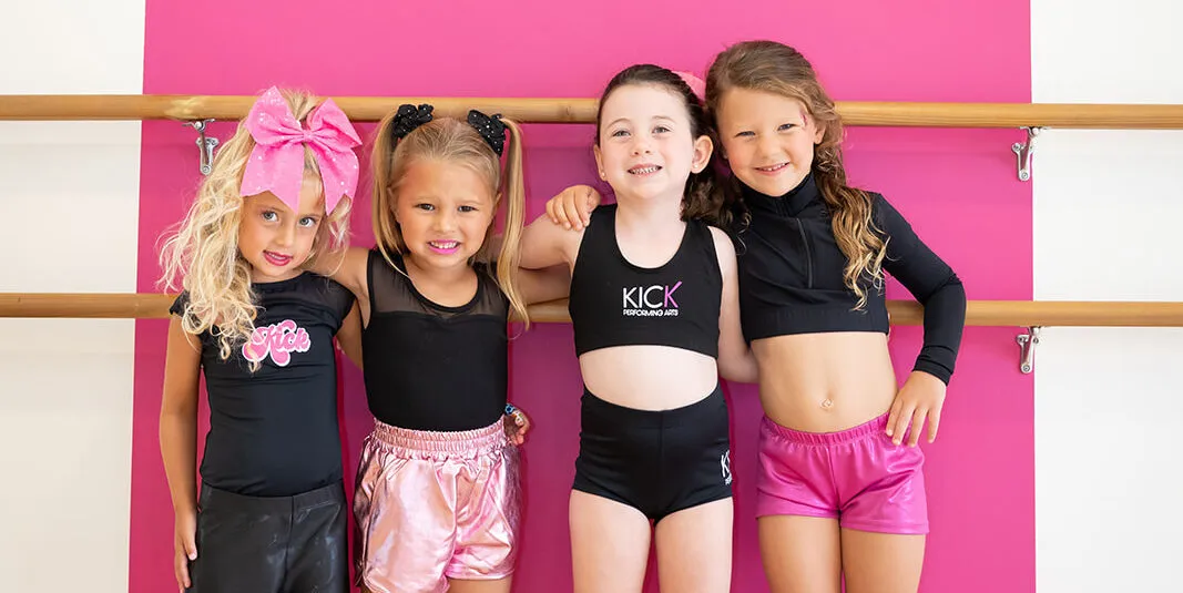 Four young KICK dancers smiling together in front of the pink studio wall
